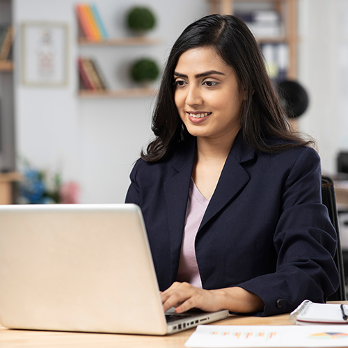 woman in a black jacket sitting at a desk typing on a laptop